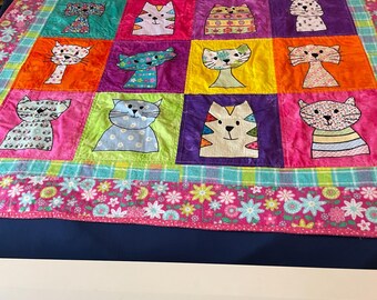 May include: A colorful patchwork quilt featuring a grid of sixteen square blocks, each displaying a unique cat illustration in various patterns and bright colors. The quilt is bordered by a vibrant pink fabric with a floral print and a plaid trim.