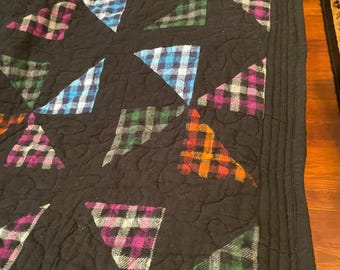 May include: A black quilt featuring a pattern of triangular patches made from multicolored plaid flannel fabric. The quilt has a solid black border with decorative stitching and a repeating quilted texture across the entire surface.