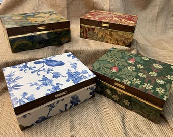 Puede incluir: Cuatro cajas rectangulares de madera con tapas abatibles, cada una con distintos patrones decorativos. Una caja tiene un estampado de pájaros y flores en azul y blanco, otra un diseño floral verde, una un patrón floral rojo y otra un estampado floral oscuro.