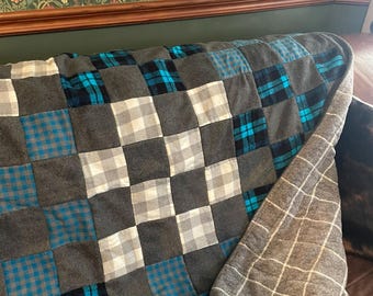 May include: A patchwork quilt constructed from square fabric pieces in shades of charcoal gray, white, and bright blue. The quilt features a mix of solid gray squares and plaid patterns in blue and white, with a gray and white grid backing visible.