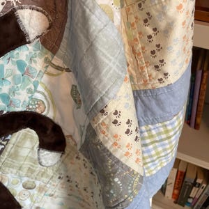 Puede incluir: Una colcha de retazos con varios cuadrados de tela en tonos azul, verde y crema. El dise&ntilde;o incluye patrones de huellas, c&iacute;rculos y cuadros. Formas de tela de felpa marr&oacute;n oscuro est&aacute;n aplicadas sobre la superficie de la colcha.