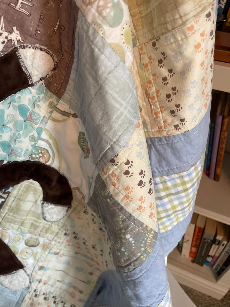 Puede incluir: Una colcha de retazos con varios cuadrados de tela en tonos azul, verde y crema. El dise&ntilde;o incluye patrones de huellas, c&iacute;rculos y cuadros. Formas de tela de felpa marr&oacute;n oscuro est&aacute;n aplicadas sobre la superficie de la colcha.