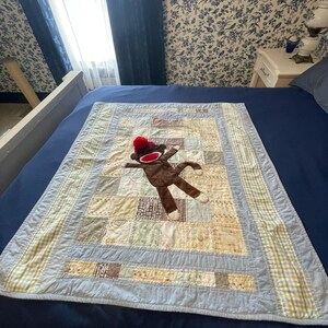 Puede incluir: Una colcha de retazos rectangular con una variedad de cuadrados de tela estampados en azul claro, beige y blanco. Un mu&ntilde;eco de mono hecho de calcet&iacute;n marr&oacute;n con un gorro rojo y boca blanca est&aacute; colocado en el centro de la colcha con borde azul.
