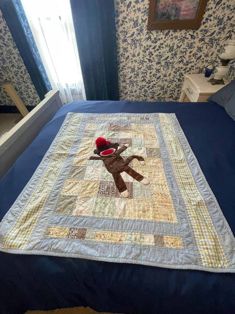Puede incluir: Una colcha de retazos rectangular con una variedad de cuadrados de tela estampados en azul claro, beige y blanco. Un mu&ntilde;eco de mono hecho de calcet&iacute;n marr&oacute;n con un gorro rojo y boca blanca est&aacute; colocado en el centro de la colcha con borde azul.