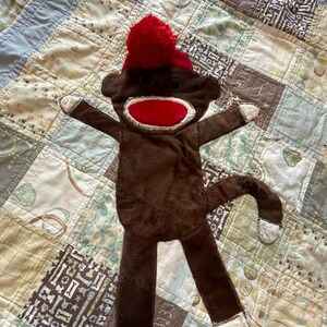 Puede incluir: Un juguete de mono de peluche marr&oacute;n con boca roja, ojos de bot&oacute;n negros y un gorro rojo con un pomp&oacute;n rojo en la parte superior. El mono tiene detalles de tela blanca en las manos, los pies y la punta de la cola, con un cuerpo plano y suave.