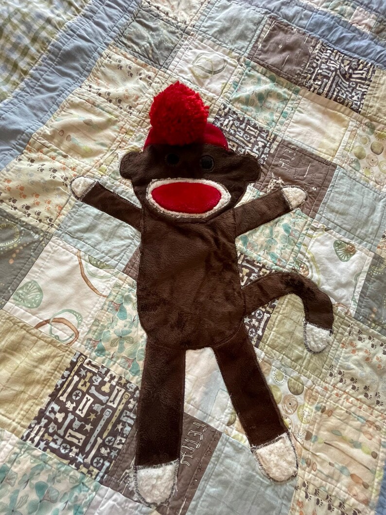 Puede incluir: Un juguete de mono de peluche marr&oacute;n con boca roja, ojos de bot&oacute;n negros y un gorro rojo con un pomp&oacute;n rojo en la parte superior. El mono tiene detalles de tela blanca en las manos, los pies y la punta de la cola, con un cuerpo plano y suave.