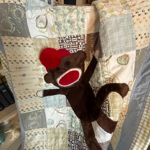 Puede incluir: Una colcha de retazos con varios cuadrados que presentan patrones de huellas, huesos y formas abstractas en tonos azules, verdes y marrones. Un peluche de mono hecho de calcet&iacute;n marr&oacute;n con un gorro y una boca rojos se encuentra sobre la colcha.