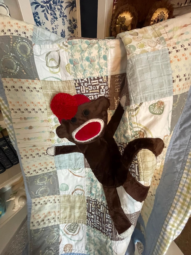 Puede incluir: Una colcha de retazos con varios cuadrados que presentan patrones de huellas, huesos y formas abstractas en tonos azules, verdes y marrones. Un peluche de mono hecho de calcet&iacute;n marr&oacute;n con un gorro y una boca rojos se encuentra sobre la colcha.