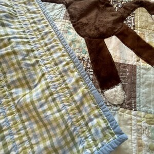Puede incluir: Un juguete de peluche de mono marr&oacute;n con boca roja y detalles blancos en manos y pies, sobre una colcha de retazos con tela de cuadros vichy verde y blanca, cuadrados marrones con la palabra FETCH y varios patrones en azul claro y blanco.