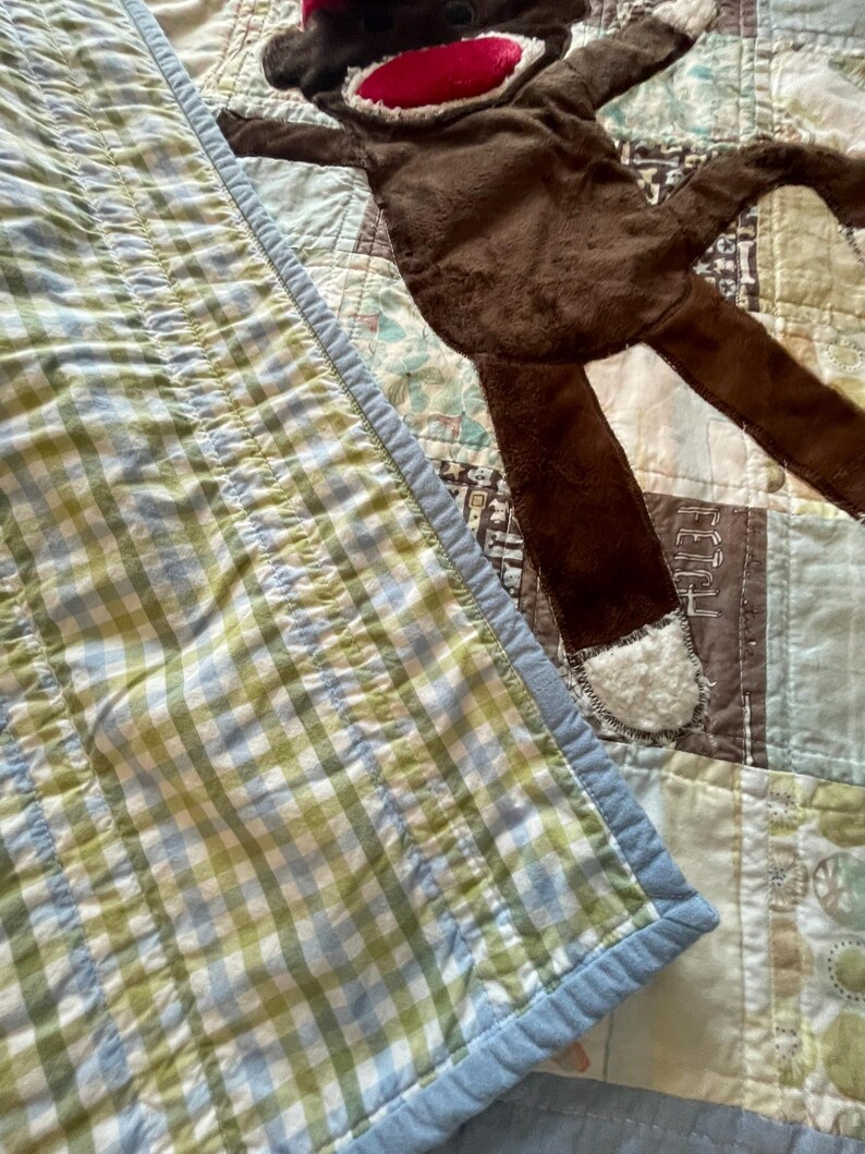 Puede incluir: Un juguete de peluche de mono marr&oacute;n con boca roja y detalles blancos en manos y pies, sobre una colcha de retazos con tela de cuadros vichy verde y blanca, cuadrados marrones con la palabra FETCH y varios patrones en azul claro y blanco.