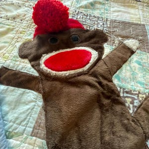 Puede incluir: Un juguete de peluche de mono hecho de calcet&iacute;n marr&oacute;n con un gorro de pomp&oacute;n rojo brillante, una boca ovalada roja grande con costuras blancas y dos ojos circulares oscuros. El juguete tiene brazos extendidos con manos blancas.