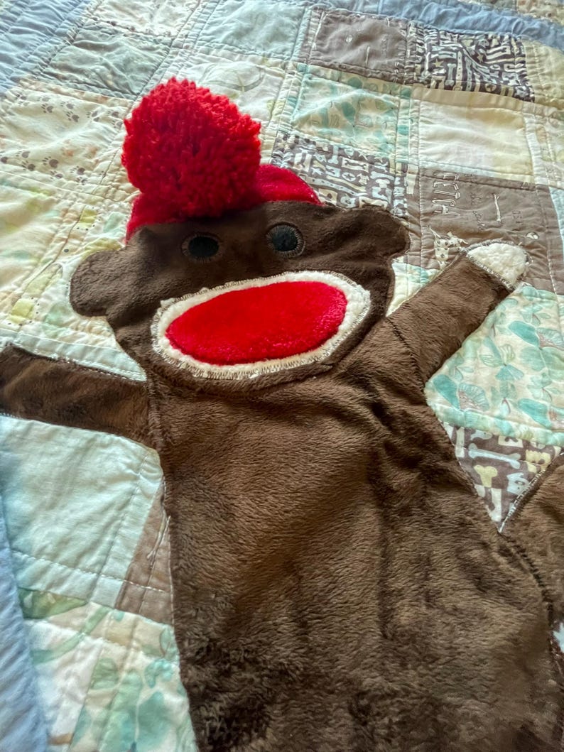 Puede incluir: Un juguete de peluche de mono hecho de calcet&iacute;n marr&oacute;n con un gorro de pomp&oacute;n rojo brillante, una boca ovalada roja grande con costuras blancas y dos ojos circulares oscuros. El juguete tiene brazos extendidos con manos blancas.