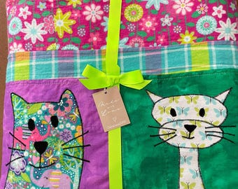 May include: A folded quilt featuring a top border of pink fabric with a colourful floral pattern, a middle plaid strip, and a bottom section with two cat appliqués on purple and green backgrounds. A bright neon green ribbon ties the quilt.