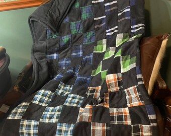 May include: A patchwork quilt constructed from square fabric pieces in various plaid patterns, including green, blue, orange, and white, interspersed with solid black squares. The quilt features a dark black border and a soft, textured surface.