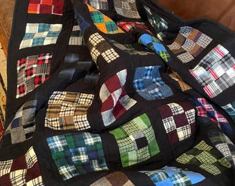 May include: A patchwork quilt constructed from various plaid and checkered fabric squares in colors including red, blue, green, yellow, and white. The squares are separated by solid black fabric borders, creating a grid pattern across the entire blanket.