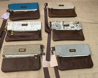 May include: Six rectangular fold-over clutch bags with brown leather-like bases and varied top panels in blue, beige, white with 'Live Laugh Love' text, coffee-themed print, woven tan, and grey fabric, each featuring a wrist strap and metal hardware.