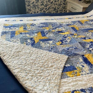May include: A quilted bedspread featuring a patchwork design of blue, white, and yellow fabric squares. The pattern includes star motifs and various floral and geometric prints. The underside of the quilt is white with a subtle blue floral pattern.