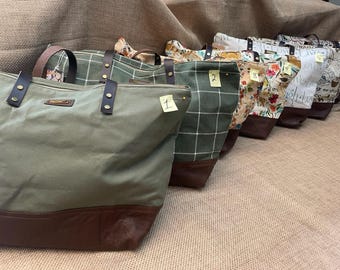 May include: A row of six tote bags with brown leather bases and various fabric patterns, including solid olive green, green plaid, floral prints, and text-based designs. Each bag features dark brown leather handles secured with brass-colored rivets.