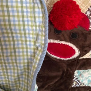 Puede incluir: Un juguete de peluche de mono marr&oacute;n con un pomp&oacute;n de lana rojo brillante en la cabeza y una boca roja, parcialmente cubierto por una manta de tela de cuadros vichy azul claro, verde y blanco con borde azul claro, sobre un textil acolchado.