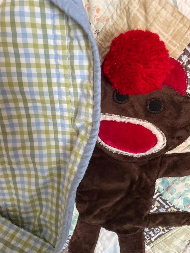 Puede incluir: Un juguete de peluche de mono marr&oacute;n con un pomp&oacute;n de lana rojo brillante en la cabeza y una boca roja, parcialmente cubierto por una manta de tela de cuadros vichy azul claro, verde y blanco con borde azul claro, sobre un textil acolchado.