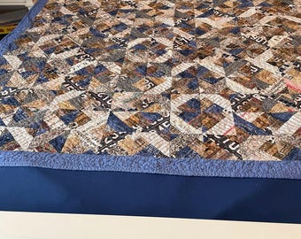 May include: A patchwork quilt featuring a repeating geometric pattern of triangles in shades of blue, brown, tan, and white. The fabric includes printed text, numbers, and map-like graphics, finished with a solid blue border and quilted texture.