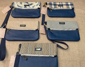May include: Five rectangular fold-over clutch bags with navy blue leather-textured lower sections and silver-tone zippers. Each bag features a unique upper fabric panel in floral, plaid, tan, grey, or woven patterns, and includes a navy wrist strap.
