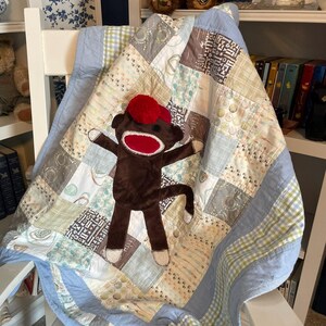 Puede incluir: Una colcha de retazos con cuadrados de tela estampados en azul claro, crema y marr&oacute;n con un borde azul claro s&oacute;lido. Un peluche de mono hecho de calcet&iacute;n marr&oacute;n con gorro y boca rojos est&aacute; centrado sobre la superficie de la colcha.