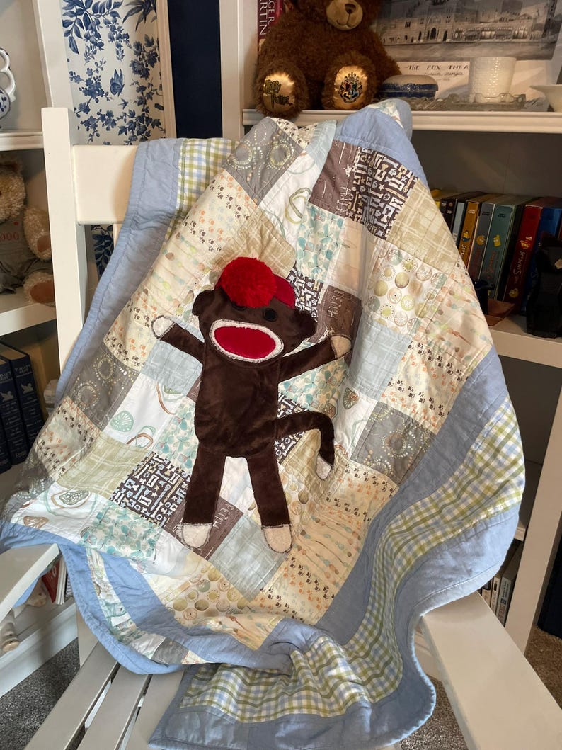 Puede incluir: Una colcha de retazos con cuadrados de tela estampados en azul claro, crema y marr&oacute;n con un borde azul claro s&oacute;lido. Un peluche de mono hecho de calcet&iacute;n marr&oacute;n con gorro y boca rojos est&aacute; centrado sobre la superficie de la colcha.