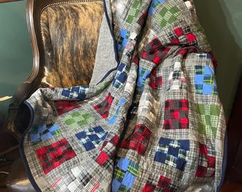 May include: A patchwork quilt constructed from various flannel fabric squares in shades of gray, red, green, and blue. The quilt features a repeating plaid pattern throughout, finished with a solid dark navy blue fabric binding along the edges.