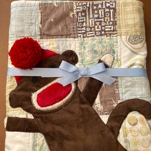 Puede incluir: Una colcha de retazos con una aplicaci&oacute;n de mono de peluche marr&oacute;n que lleva un gorro con pomp&oacute;n rojo y tiene una boca de color rojo brillante. La colcha est&aacute; atada con una cinta azul claro y muestra varios patrones de huesos y c&iacute;rculos.