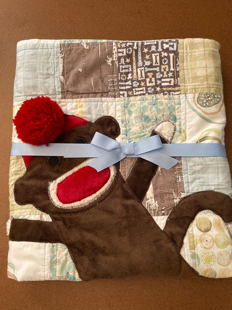 Puede incluir: Una colcha de retazos con una aplicaci&oacute;n de mono de peluche marr&oacute;n que lleva un gorro con pomp&oacute;n rojo y tiene una boca de color rojo brillante. La colcha est&aacute; atada con una cinta azul claro y muestra varios patrones de huesos y c&iacute;rculos.