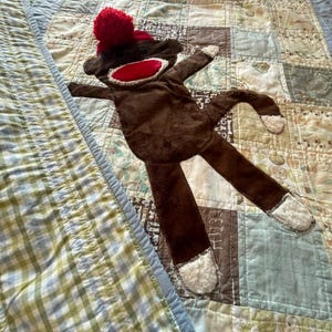 Puede incluir: Un juguete de peluche con forma de mono hecho de calcet&iacute;n, con cuerpo marr&oacute;n oscuro, boca roja brillante y un pomp&oacute;n rojo en la cabeza. El mono tiene tela blanca texturizada en manos y pies, ojos de bot&oacute;n negros y una cola larga.