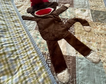 May include: A plush sock monkey toy featuring a dark brown body, a bright red mouth, and a red pom-pom on its head. The monkey has white textured fabric on its hands and feet, with black button eyes and a long tail, lying flat on a quilt.