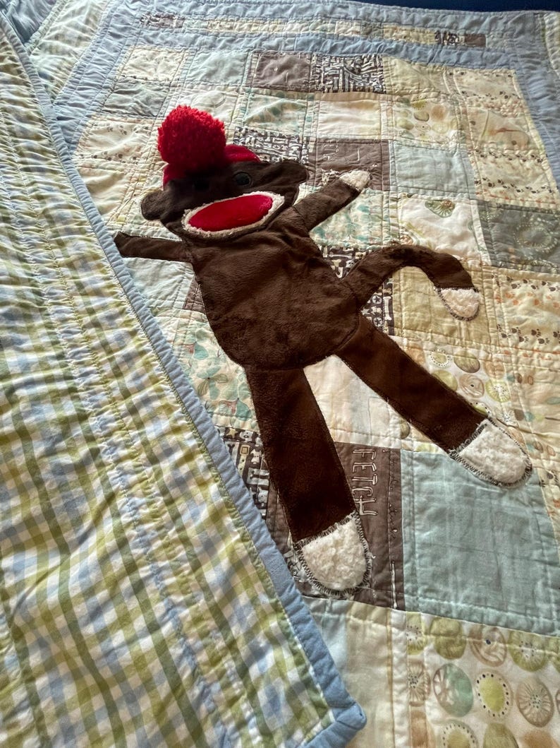 Puede incluir: Un juguete de peluche con forma de mono hecho de calcet&iacute;n, con cuerpo marr&oacute;n oscuro, boca roja brillante y un pomp&oacute;n rojo en la cabeza. El mono tiene tela blanca texturizada en manos y pies, ojos de bot&oacute;n negros y una cola larga.