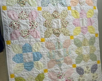 May include: A handmade quilt featuring a grid of twelve floral and patterned fabric blocks in various pastel colors. Each block consists of four petal-shaped pieces surrounding a white square center, separated by small yellow corner squares.