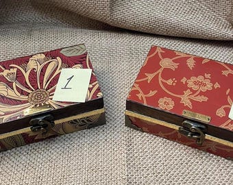 May include: Two rectangular wooden boxes with metal latches and gold-toned trim. Box 1 features a dark red and cream floral pattern. Box 2 has a red surface with a gold-toned floral vine design. Each box has a small paper label marked 1 or 2.