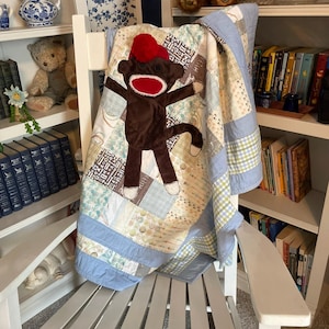 May include: A patchwork quilt featuring a brown sock monkey applique with a red pom-pom hat and a red mouth. The quilt is constructed from various light blue, cream, and patterned fabric squares, finished with a solid light blue border trim.