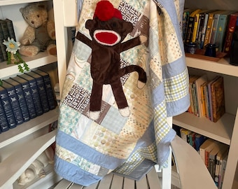 May include: A patchwork quilt featuring a brown sock monkey applique with a red pom-pom hat and a red mouth. The quilt is constructed from various light blue, cream, and patterned fabric squares, finished with a solid light blue border trim.