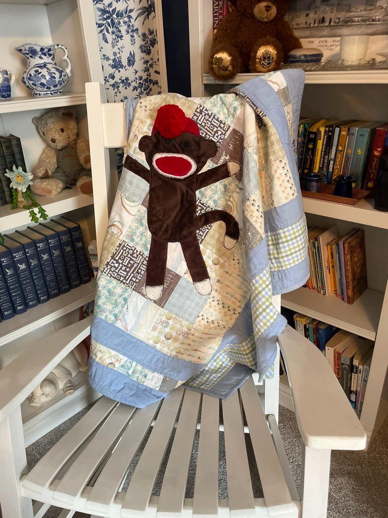 Puede incluir: Una colcha de retazos con una aplicaci&oacute;n de mono de calcet&iacute;n marr&oacute;n con un gorro de pomp&oacute;n rojo y una boca roja. La colcha est&aacute; hecha de varios cuadrados de tela de color azul claro, crema y estampados, con un borde de color azul claro.