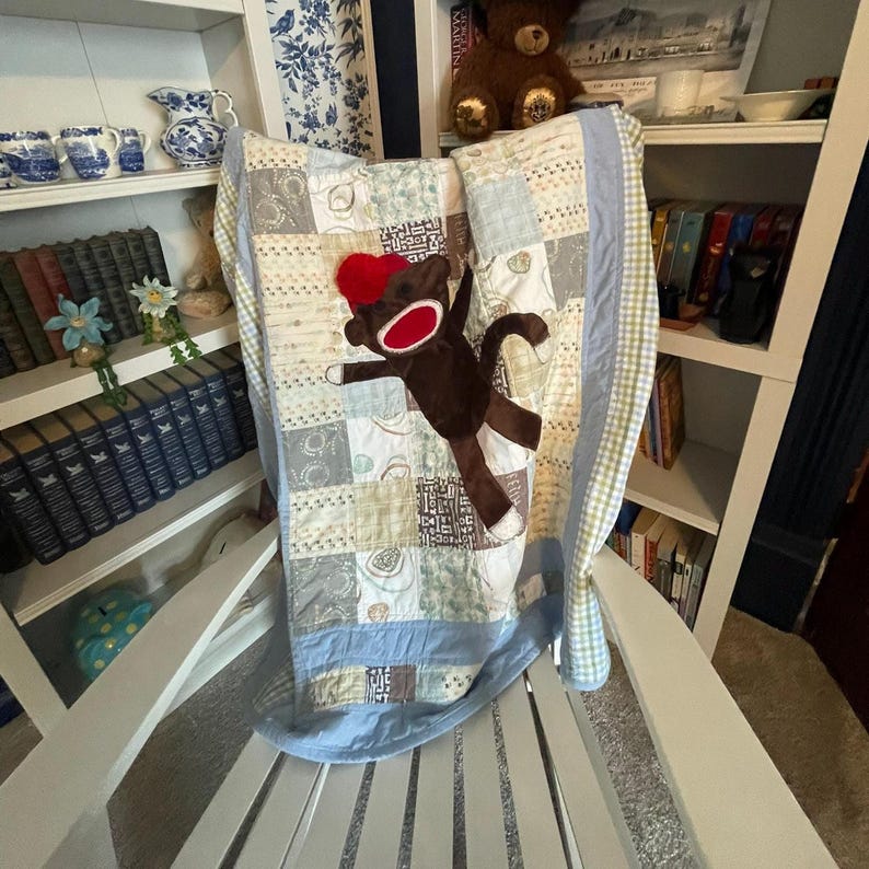 Puede incluir: Una colcha de retazos con varios cuadrados de tela estampada en azul claro, crema y marr&oacute;n. Un peluche de mono hecho de calcet&iacute;n marr&oacute;n con boca roja y gorro rojo est&aacute; sujeto al centro de la colcha, que tiene un borde de color azul claro.