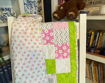 May include: A patchwork quilt featuring a mix of white, pink, and lime green fabrics. The design includes squares with floral patterns, grey chevron stripes, and solid colours. The quilt is draped over a white wooden chair, showcasing a quilted texture.