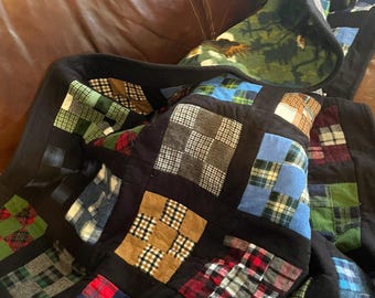 May include: A patchwork quilt constructed from various squares of plaid and flannel fabric in shades of red, blue, green, brown, and black. The squares are separated by solid black fabric borders, and the underside features a green forest print.