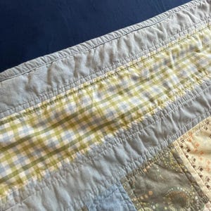 Puede incluir: Una colcha de retazos con una variedad de cuadrados de tela en tonos azul claro, verde salvia y crema. El dise&ntilde;o incluye un borde de cuadros vichy verde y azul, paneles de color azul claro s&oacute;lido y cuadrados con peque&ntilde;os patrones de huellas.