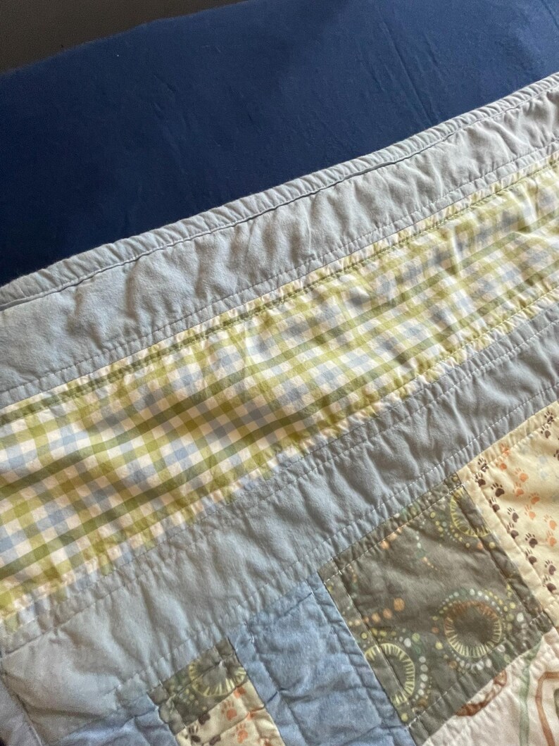 Puede incluir: Una colcha de retazos con una variedad de cuadrados de tela en tonos azul claro, verde salvia y crema. El dise&ntilde;o incluye un borde de cuadros vichy verde y azul, paneles de color azul claro s&oacute;lido y cuadrados con peque&ntilde;os patrones de huellas.
