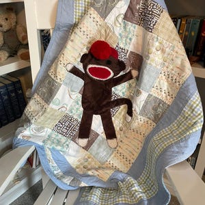 Puede incluir: Una colcha de retazos con cuadrados de tela en azul claro, beige y blanco con diversos patrones, incluyendo rayas, cuadros y motivos peque&ntilde;os. Un peluche de mono hecho de calcet&iacute;n marr&oacute;n con un gorro de pomp&oacute;n rojo est&aacute; centrado.