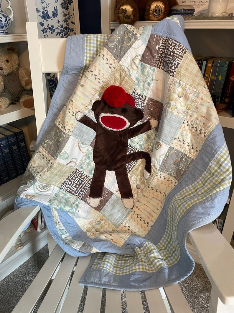 Puede incluir: Una colcha de retazos con cuadrados de tela en azul claro, beige y blanco con diversos patrones, incluyendo rayas, cuadros y motivos peque&ntilde;os. Un peluche de mono hecho de calcet&iacute;n marr&oacute;n con un gorro de pomp&oacute;n rojo est&aacute; centrado.