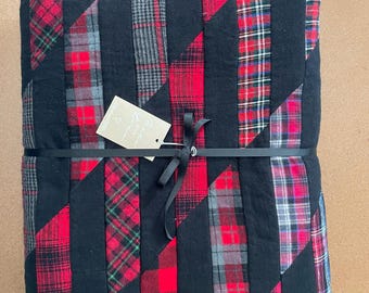 May include: A folded quilt featuring a patchwork design of solid black fabric strips alternating with various red, black, and gray plaid patterns. A black ribbon is tied around the center, holding a beige tag with the text Made with love.