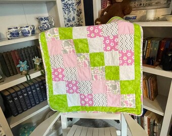 Könnte beinhalten: Eine quadratische Patchwork-Decke mit einem sich wiederholenden Muster aus hellgrünen, weißen und rosafarbenen Stoffquadraten. Die Decke enthält verschiedene Drucke wie kleine weiße Blumen, graue Zickzackstreifen und rosa Vichy-Karos, eingefasst mit einem hellgrünen Rand.