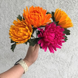 May include: A bouquet of handmade felt flowers in vibrant colors. The arrangement features orange, yellow, and fuchsia chrysanthemums with green leaves. The flowers are held in a hand, showcasing the detailed craftsmanship of the floral arrangement.