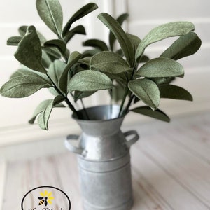 May include: A galvanized metal milk jug vase holds a bundle of sage green felt leaves. The leaves have a textured appearance and are arranged to create a full, natural look. The vase has two small handles and a slightly distressed finish. The logo for ShellBee's Crafts and More is in the lower left corner.